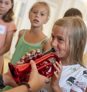 Photo: A young girl plays on a small trumpet in red color with the support of another person, of which only their hands are visible, holding the trumpet along. In the background there are other interested girls.
