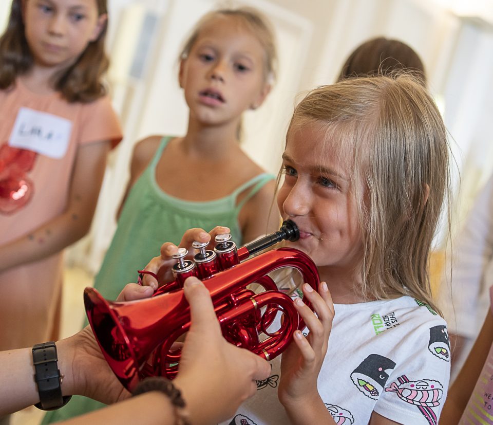Foto: Ein junges Mädchen spielt auf einer kleinen Trompete in roter Farbe mit Unterstützung einer anderen Person, von der nur deren Hände sichtbar sind, welche die Trompete mithalten. Im Hintergrund befinden sich weitere interessierte Mädchen.