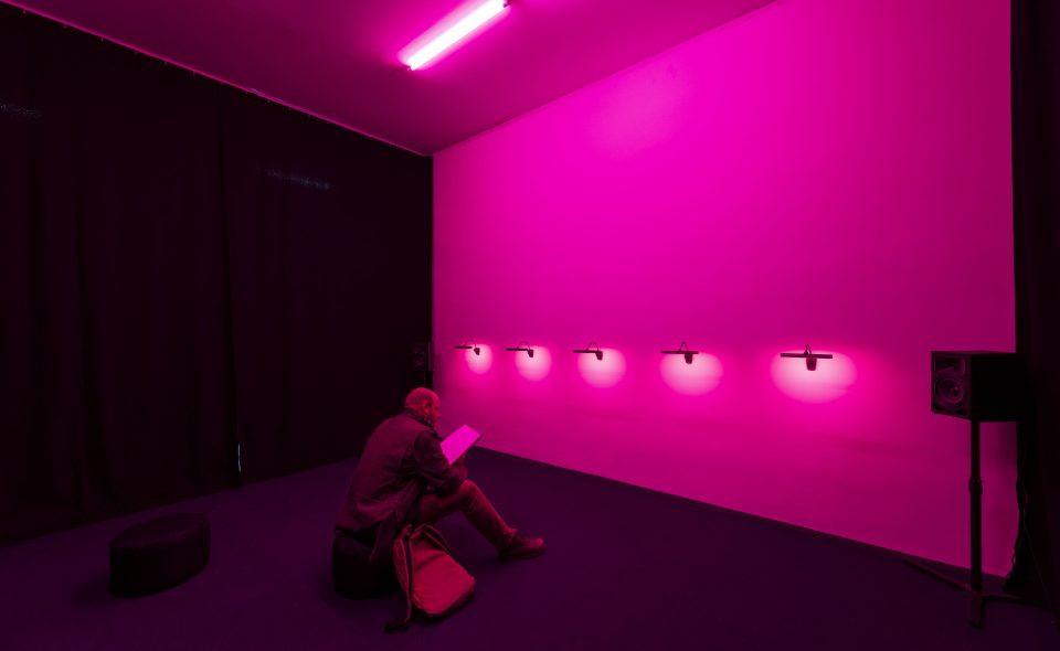 Foto von einem Mann, der auf einem Hocker in einem leeren, pink beleuchteten Raum sitzt.