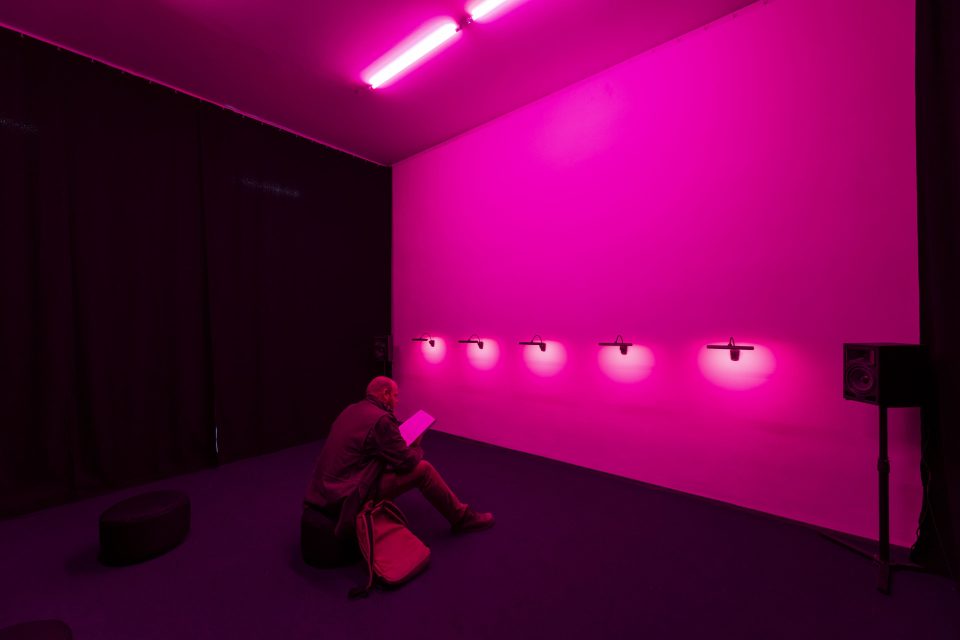 Foto von einem Mann, der auf einem Hocker in einem leeren, pink beleuchteten Raum sitzt.