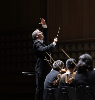 Markus Poschner beim Dirigieren des Bruckner Orchester Linz, von welchem wenige Musiker:innen von hinten zu sehen sind.