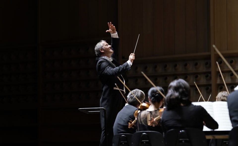Markus Poschner beim Dirigieren des Bruckner Orchester Linz, von welchem wenige Musiker:innen von hinten zu sehen sind.