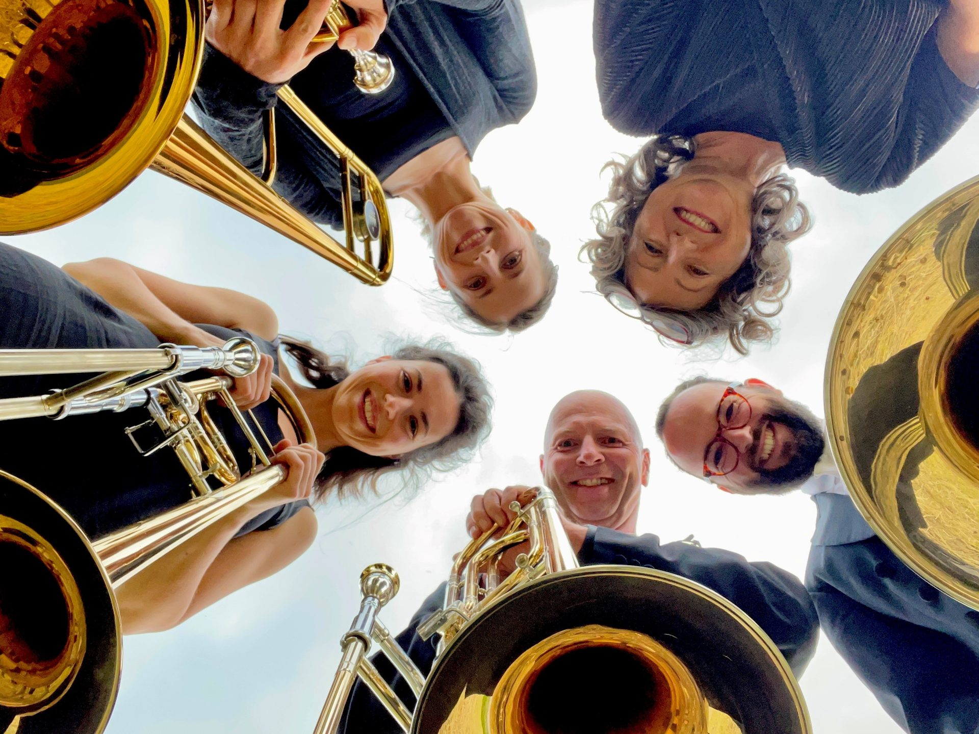 Musicians stand in a circle with their instruments. They laugh into the camera in the center of the circle.