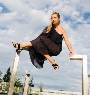 Photo of Silke Grabinger, dressed in black, poses acrobatically in front of a clear sky. Here she supports herself on an iron scaffold.