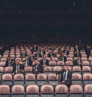 Concert hall, facing the audience, the music group Vienna Brass Connection consisting of twenty-one men sits spread out in the room and each has a musical instrument in his hand